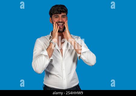 Homme d'affaires avec chemise blanche et cheveux foncés tenant le visage dans l'étonnement, exprimant l'excitation et la surprise sur fond bleu Banque D'Images