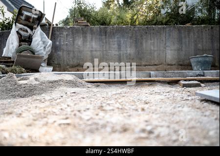 Une scène de travail de sol avec du sable, des blocs de béton et des outils contre un mur de béton, mis en place pour un projet de pavage ou d'aménagement paysager. Banque D'Images