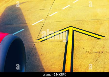 Flèche directionnelle jaune sur une piste d'aéroport, photographiée depuis une fenêtre d'avion avec un moteur rose en vue, symbolisant le mouvement, le voyage et l'étoile Banque D'Images