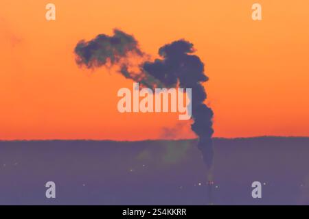 Un coucher de soleil saisissant présente un panache de fumée sombre magnifiquement silhouette sur un ciel orange vibrant Banque D'Images