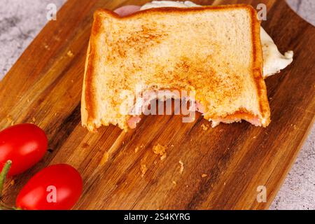 Un délicieux sandwich à moitié mangé d'oeuf, de jambon et de fromage est empilé verticalement, accompagné de tomates cerises fraîches sur une table en béton gris Banque D'Images