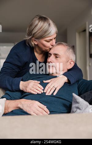 Un couple d'aînés partage un moment tendre sur le canapé la femme embrasse amoureusement la tête de l'homme cette scène rayonne de chaleur, d'affection et une émotion profonde Banque D'Images