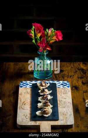 Élégants canapés de craquelins surmontés de ragoût satay de légumes et d'arachides, servis sur une ardoise noire sur une planche de bois Un vase avec des fleurs rouges ajoute un vibra Banque D'Images