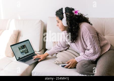 Une jeune femme latine se détend sur un canapé confortable à la maison, portant des écouteurs et parcourant son ordinateur portable elle tient un téléphone, profitant de son temps libre dans une comfo Banque D'Images