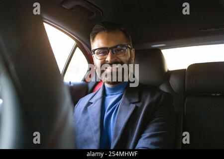 Homme d'affaires indien souriant portant des lunettes et des vêtements d'hiver assis dans une voiture, regardant directement la caméra. Banque D'Images