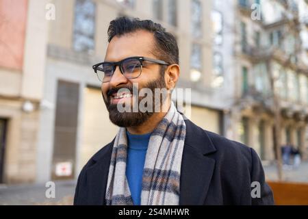 Un homme indien joyeux en tenue d'hiver, comprenant une écharpe et un manteau, des lunettes de sport et une barbe, se prélassant au soleil dans une rue urbaine, regardant loin Banque D'Images