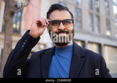 Un homme d'affaires indien en tenue d'hiver élégante, ajustant ses lunettes tout en souriant en toute confiance à l'extérieur, près d'un fond urbain. Banque D'Images