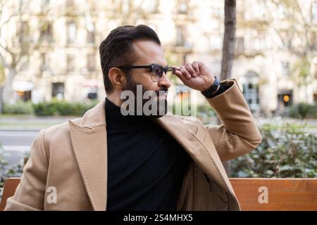 Élégant homme indien portant des lunettes, un col roulé, et un manteau beige ajuste ses lunettes lorsqu'il est assis sur un banc dans une ville pendant l'hiver. Banque D'Images