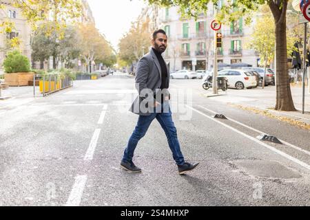 Un homme d'affaires indien traverse une rue de la ville, vêtu d'une élégante tenue d'hiver composée d'un blazer gris, d'un pull à col tortue noir et d'un Jean bleu Banque D'Images