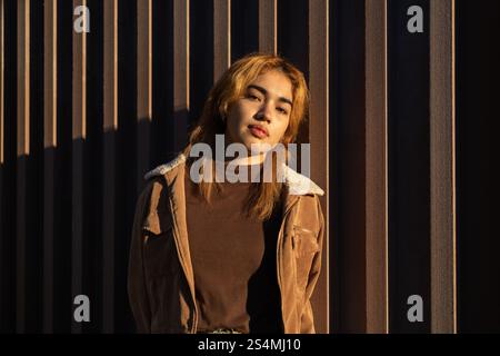 Une femme de race mixte s'appuie contre un mur texturé, baignée de lumière chaude du soleil sa tenue décontractée et son expression confiante créent un vi harmonieux et moderne Banque D'Images