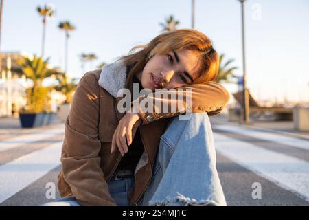 Une femme de race mixte vêtue de façon décontractée est assise sur un passage piétonnier sur une promenade, entourée de palmiers et d'une douce lumière du soir, créant un a serein Banque D'Images