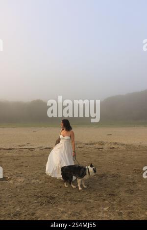 Une femme dans une robe blanche fluide marche avec son chien sur une plage sereine le fond est enveloppé dans la brume, créant une atmosphère tranquille et éthéré Banque D'Images