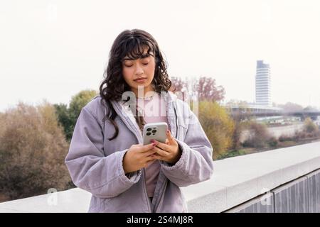 Jeune femme latine dans une veste décontractée vérifiant son téléphone à l'extérieur, debout sur un pont avec des arbres et un paysage urbain en arrière-plan détendu et pourtant Banque D'Images