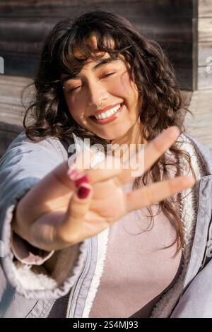 Une jeune femme latine dynamique profite d'une journée ensoleillée, clignotant un signe de paix avec un sourire joyeux. Son ambiance détendue et insouciante incarne le bonheur et la positivité Banque D'Images