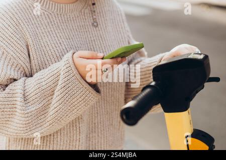 Femme recadrée méconnaissable scanne le code QR avec un smartphone pour louer un scooter électrique. L'accent est mis sur ses mains tenant à la fois le téléphone et le scooter Banque D'Images