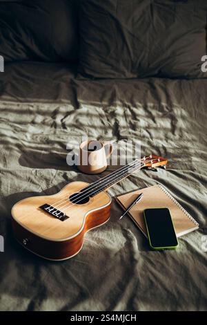 Un cadre confortable et créatif avec un ukulélé, une tasse en céramique, un carnet ouvert avec un stylo et un téléphone intelligent, le tout placé sur une pré de lit douce et froissée Banque D'Images
