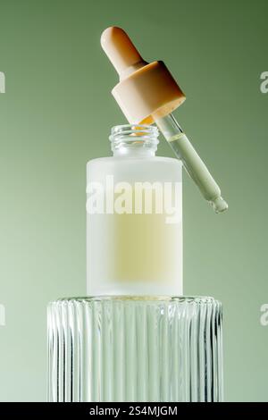 Un élégant flacon compte-gouttes de sérum avec une pipette reposant sur son bord, rempli d'un sérum laiteux, placé sur un fond vert doux. Banque D'Images