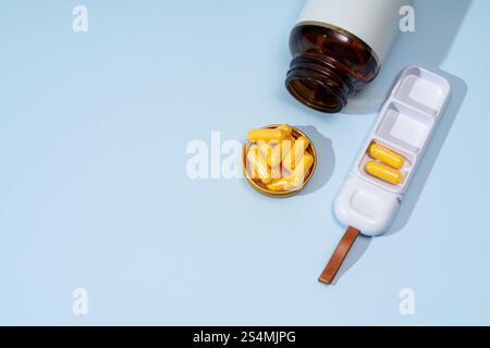 D'en haut, une image montrant une capsule jaune renversée d'une bouteille brun foncé, un organisateur de pilules avec des capsules, et un couvercle ouvert sur un fond bleu. Banque D'Images
