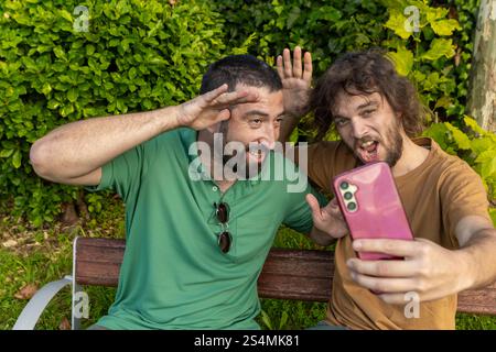 Deux amis s'amusent à prendre un selfie ludique assis sur un banc de parc l'homme en vert, qui est aveugle, sourit et pose avec enthousiasme aux côtés Banque D'Images