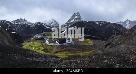 Une vue saisissante sur les montagnes volcaniques noires partiellement recouvertes de neige, avec une rivière sinueuse et mousse verte vibrante dans la vallée, sous un et dramatique Banque D'Images
