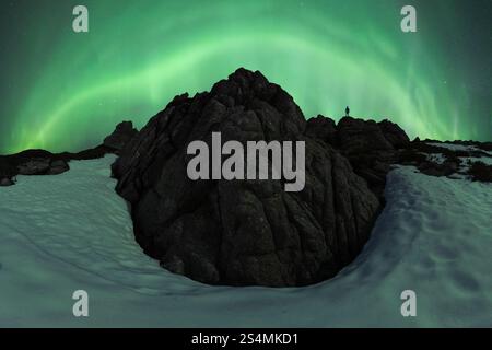 Silhouette de personne méconnaissable debout au sommet de rochers enneigés accidentés sous une aurore boréale vibrante dans une nuit nordique d'hiver sereine. Banque D'Images