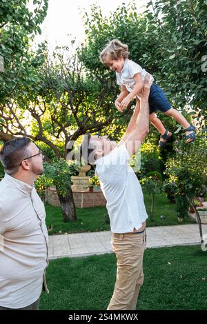 Un couple gay aimant profite du temps de qualité avec leur fils joyeux dans un jardin luxuriant. Un parent soulève l'enfant de manière ludique comme l'autre regarde wi Banque D'Images