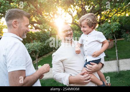 Un joyeux couple gay partage un moment tendre avec leur fils dans un jardin ensoleillé, entouré de verdure luxuriante. L'image rayonne de chaleur, d'amour et de famille Banque D'Images