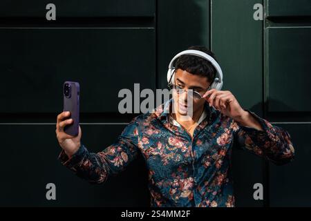 Un homme latin tendance en chemise florale et lunettes de soleil prend un selfie à l'extérieur, mettant en valeur un style moderne avec des écouteurs sur. La photo capture la confiance A. Banque D'Images