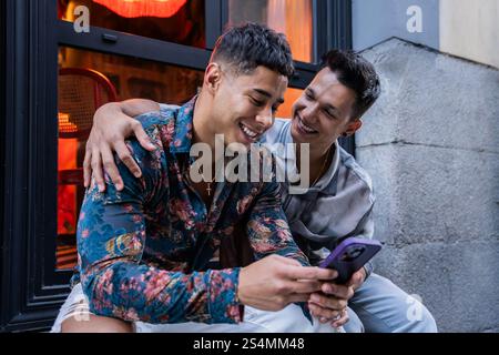 Un joyeux couple gay latin est assis devant un café, partageant un moment sur un téléphone. Ils sont souriants et embrassants, avec un décor chaleureux visible à travers la fenêtre Banque D'Images