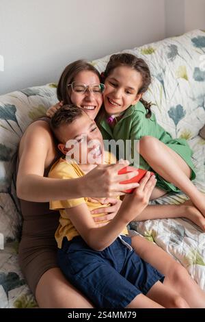 Une famille joyeuse partage un moment tendre sur le canapé. La mère aimante embrasse sa fille avec une trachéotomie et son fils avec une paralysie cérébrale, capturin Banque D'Images