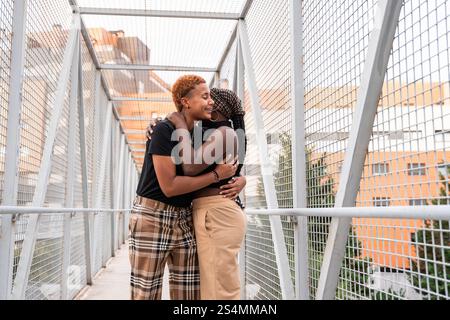 Un joyeux couple LGBT multiethnique partage une étreinte chaleureuse sur un pont de passerelle métallique, entouré de bâtiments de la ville. La scène représente l'amour, la diversité, Banque D'Images