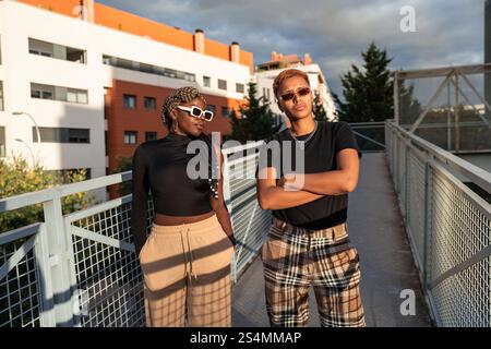 Un élégant couple LGBT multiethnique pose en toute confiance sur un pont urbain sous la lumière chaude et dorée du soleil. Les deux portent des lunettes de soleil et une tenue décontractée, expres Banque D'Images