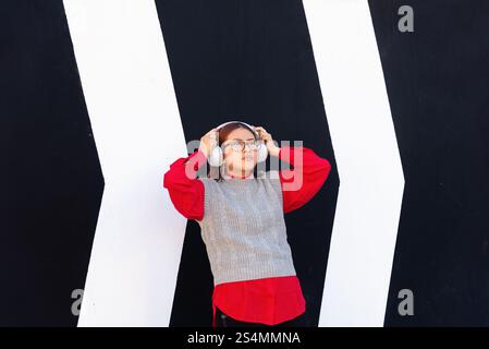 Une femme latina se tient debout devant un mur géométrique noir et blanc frappant, portant une chemise rouge et un gilet gris elle écoute de la musique à travers Banque D'Images