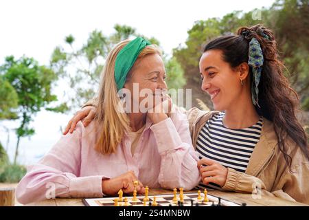 Une mère et sa fille partagent un moment joyeux en jouant aux échecs à l'extérieur, entourées de verdure et d'arbres, elles sourient chaleureusement, créant un calme et un bonheur Banque D'Images