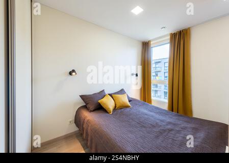 Cette chambre lumineuse et accueillante présente un décor élégant avec des rideaux moutarde, un lit confortable avec des coussins et un éclairage mural moderne. Lumière naturelle fi Banque D'Images