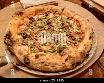 Pizza artisanale avec viandes, légumes de différentes sortes et fromage mozzarella servi sur une assiette colorée sur une table en bois Banque D'Images