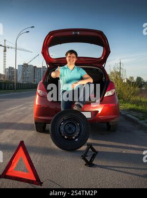 Jeune homme assis sur le coffre de la voiture et en maintenant le pied sur la roue de secours Banque D'Images