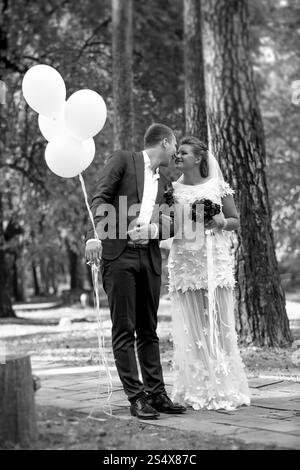 Photo en noir et blanc du marié tenant des ballons embrassant la belle mariée au parc Banque D'Images