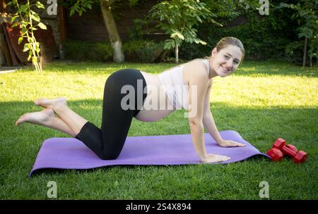 Smiling pregnant woman standing sur les genoux sur le tapis de fitness at park Banque D'Images