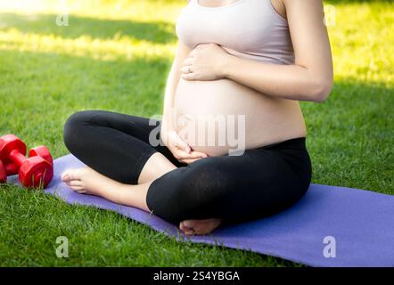 Femme enceinte active assise sur l'herbe au parc et touchant le gros ventre Banque D'Images