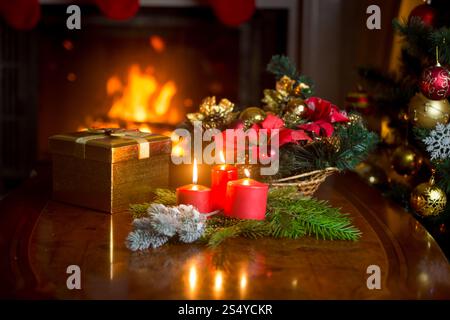 Bougies allumées, couronne de Noël et boîte cadeau dorée sur la table à côté de la cheminée allumée au salon. Image avec une faible profondeur de mise au point. Banque D'Images