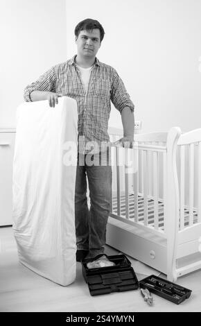 Portrait noir et blanc de jeune bel homme posant au lit bébé avec nouveau matelas Banque D'Images