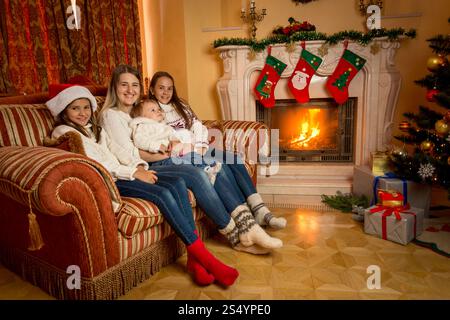 Mère assise avec ses filles sur le canapé salon à côté de la cheminée en feu à Noël Banque D'Images