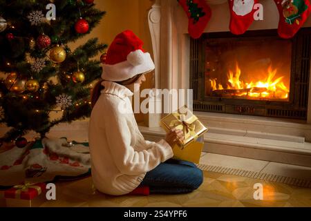Fille dans le chapeau de Père Noël assis avec boîte cadeau de Noël à la cheminée et regardant le feu Banque D'Images