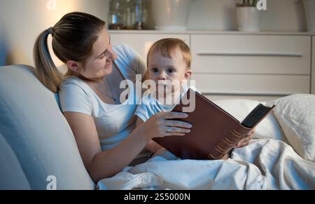 Jeune mère couchée dans le lit avec son bébé la nuit et tenant un grand livre Banque D'Images