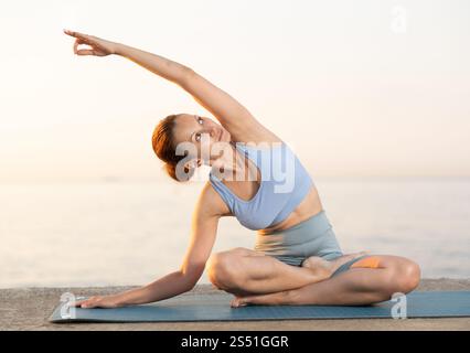 Heureuse jeune femme expérimentée professeur de fitness effectue le yoga parshvottanasana assis sur la plage le matin Banque D'Images