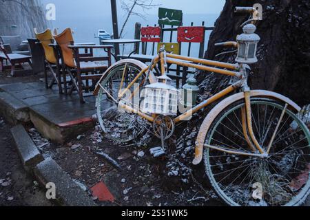 Un vélo jaune enneigé avec une lanterne repose à côté de chaises colorées et d'une table sur un patio au bord du lac pendant un matin d'hiver tranquille. Banque D'Images