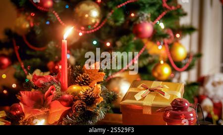 Gros plan image de couronne de Noël avec bougie allumée contre arbre de Noël coloré brillant. Gros plan photo de couronne de Noël avec bougie allumée Banque D'Images