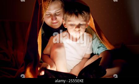 Portrait de la jeune mère couché avec son petit fils sous couverture et lui lisant le livre d'histoire. Banque D'Images