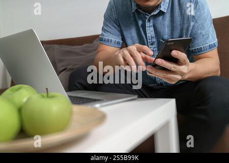 main hipster à l'aide du compte d'ordinateur portable et des paiements mobiles en ligne, omni canal, assis sur un canapé dans le salon, pommes vertes dans un plateau en bois Banque D'Images